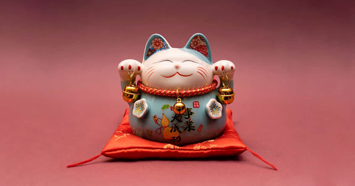 Maneki Neko sentado en un cojín con dos cascabeles en la mano